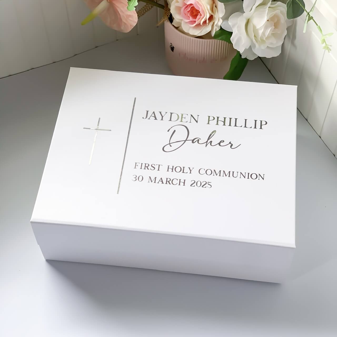 White box with personalized text for a first Holy Communion on a light surface with flowers in the background.